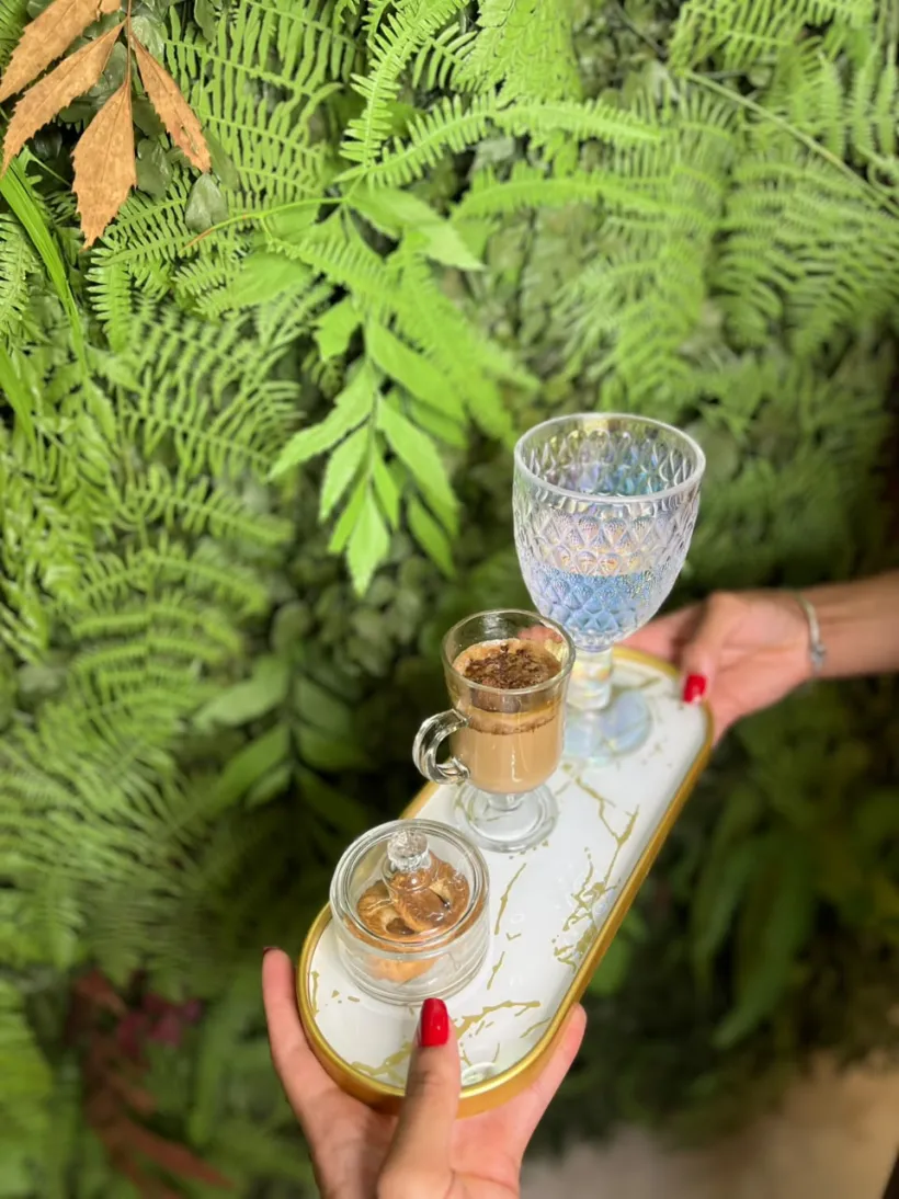 Bandeja dourada com café e taça de cristal diante da parede de plantas